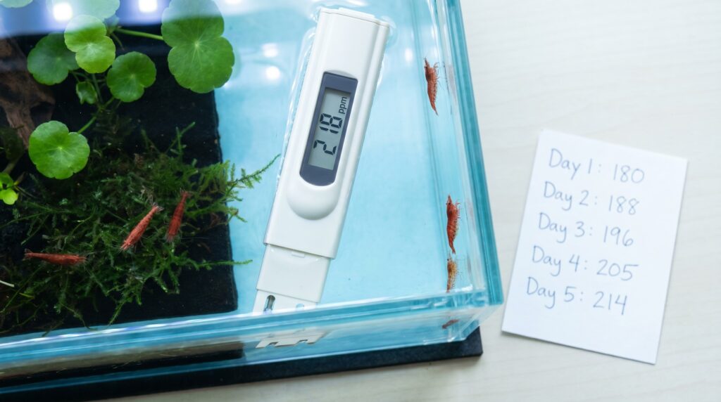 TDS meter probe in shrimp tank beside handwritten daily parameter log showing creeping TDS values